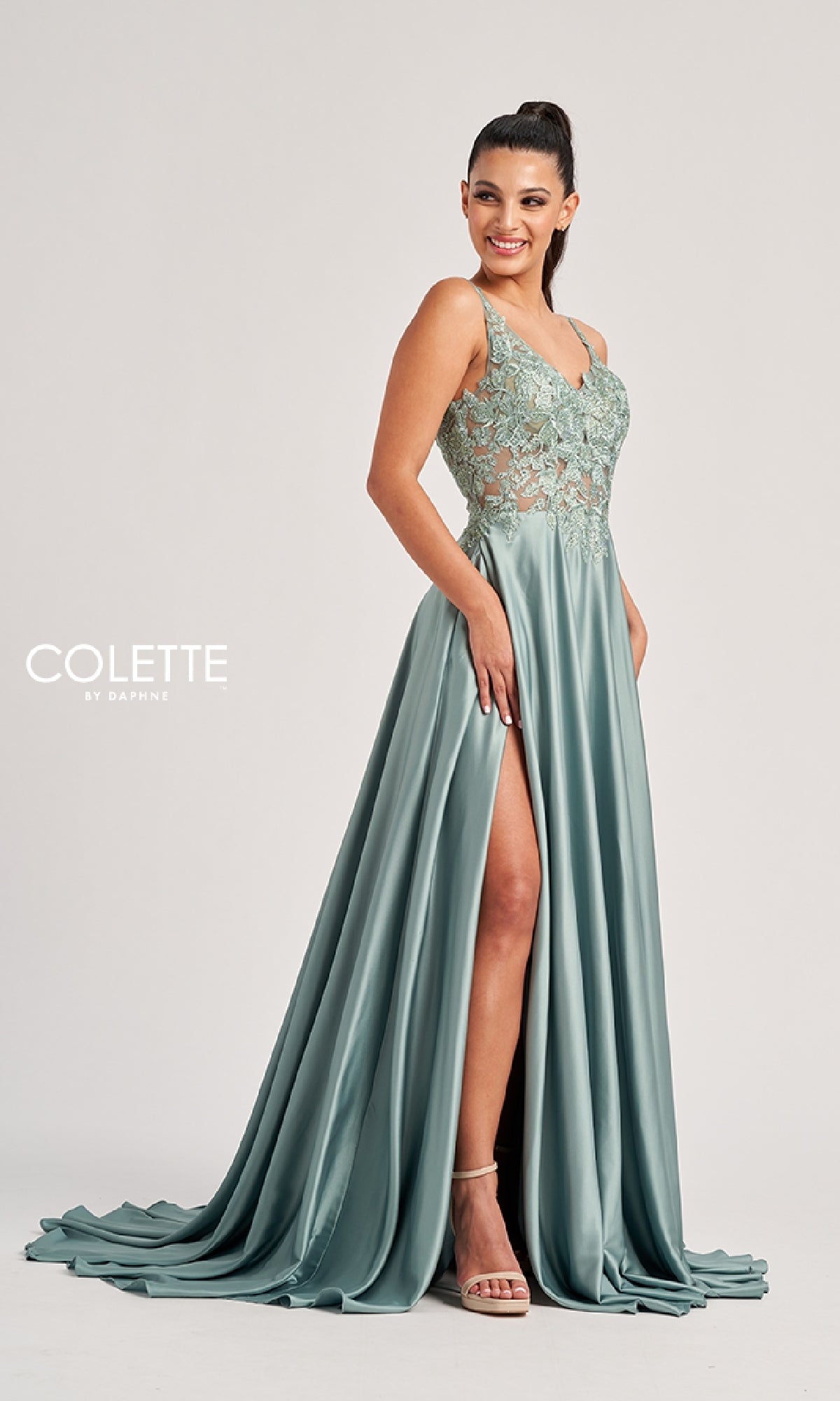 A woman stands smiling in the Colette CL8160 long prom dress, an A-line sage green gown with a lace bodice, thin straps, rhinestone details, and a high side slit.